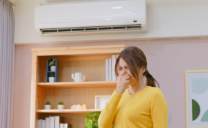 A woman in a yellow sweater covering her nose while standing near an HVAC unit, reacting to an unpleasant smell. The image highlights the importance of preparing your HVAC system for winter and addressing unusual smells when turning on the heat for the first time.