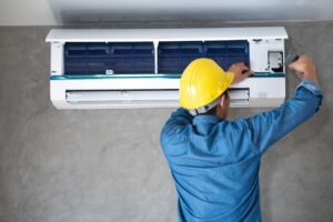AC Maintenance: A person in a yellow hard hat and blue shirt is installing or servicing a wall-mounted air conditioning unit using a screwdriver. ductless mini split repair okatie sc