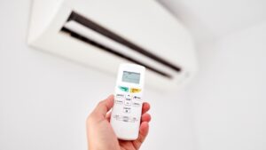 A hand holding a remote control, pointed at a wall-mounted air conditioning unit in a Hardeeville, SC home. Hvac Maintenance Plans Sun City SC
